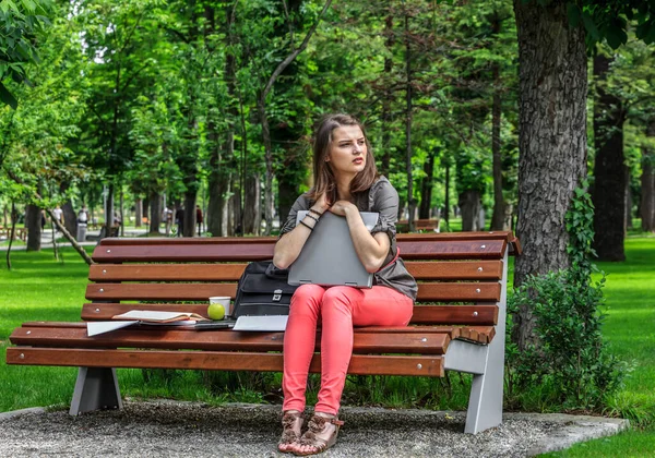 年轻的学生女人坐在公园的长椅上拥抱她的笔记本电脑 — 图库照片