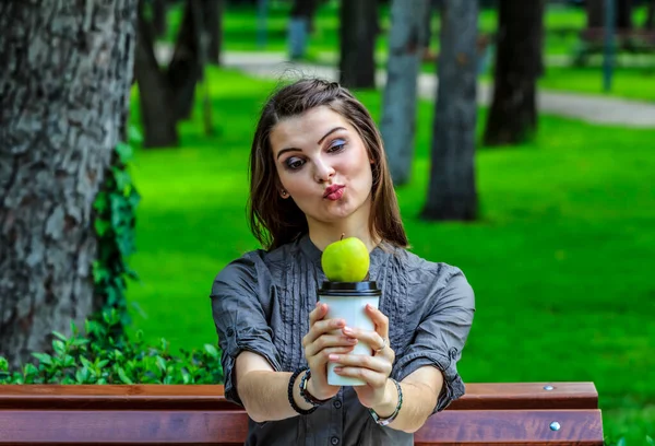 一个在公园里学习的年轻女人拿着一个绿色的苹果和一杯咖啡 望着摄像机 — 图库照片