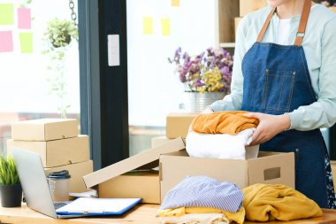 Asian female clothes shop owner folding a t-shirt and packing in a cardboard parcel box. Asian businesswoman startup entrepreneur SME owner picking up a yellow shirt before packing it in an inner box