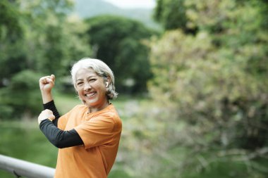 Atletik yaşlı kadın koşucu parkta koşmadan önce kollarını ve vücudunu esneterek ısınıyor..