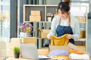 Asyalı kadın giyim mağazası sahibi tişört katlıyor ve karton bir kutuda paketliyor. Asyalı iş kadını girişimci KOBİ sahibi bir iç kutuya koymadan önce sarı bir gömlek aldı