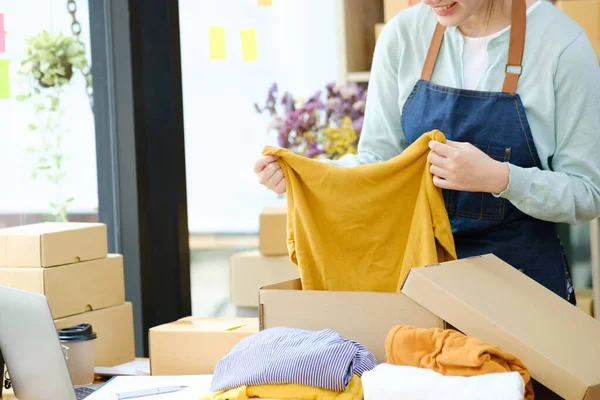 Asian female clothes shop owner folding a t-shirt and packing in a cardboard parcel box. Asian businesswoman startup entrepreneur SME owner picking up a yellow shirt before packing it in an inner box