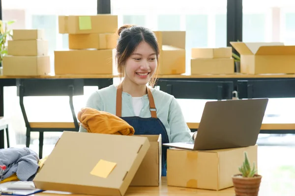 Asian female clothes shop owner folding a t-shirt and packing in a cardboard parcel box. Asian businesswoman startup entrepreneur SME owner picking up a yellow shirt before packing it in an inner box