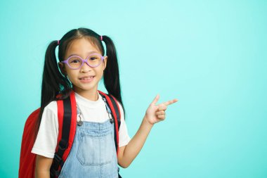 Smiling little girl in glasses pointing finger upward going to school, school concept. standing on a blue background.