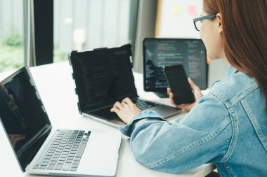 Genç programcı ya da bilişim uzmanı yaptığı işten memnun. Ofisinde çalışırken bilgisayarında ve telefonunda çalışan mutlu genç bir kadın..