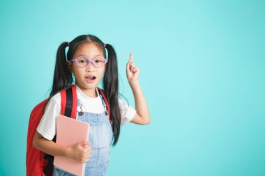 Smiling little girl in glasses pointing finger upward going to school, school concept. standing on a blue background.