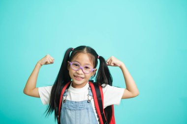 Asian little child girl raising hands up making strong gesture isolated on skyblue background. Good health concept.