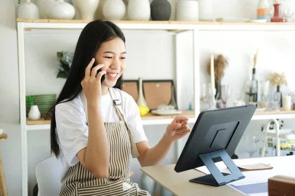若いアジアの経営者は スマートフォンやコンピュータのラップトップ上の顧客に連絡するためにラップトップ上で作業セラミック製品販売室に座っています ビジネスコンセプト — ストック写真