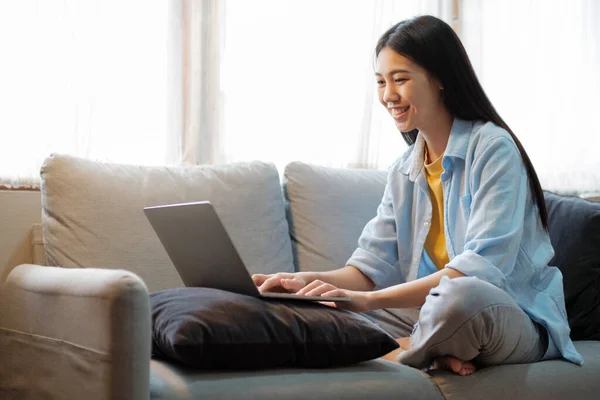 年轻的亚洲女人笑着 坐在沙发上学习或工作时使用笔记本电脑 女人在明亮的客厅舒服地坐在沙发上 一边上网学习 一边用笔记本电脑搜索信息 在家里网上学习 — 图库照片