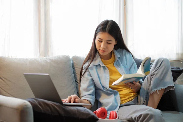 年轻的亚洲女人坐在沙发上 一边用笔记本电脑一边看书一边上网 在网上学习的年轻亚裔妇女 在线联系和在线学习概念 — 图库照片