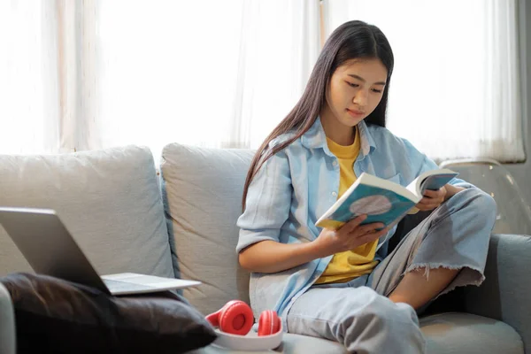年轻的亚洲女人在笔记本电脑前读书 女孩在家里看书 在沙发上呆着 教育和在线学习概念 — 图库照片