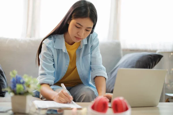 年轻的亚洲女人坐在沙发上 一边用笔记本电脑一边看书一边上网 在网上学习的年轻亚裔妇女 在线联系和在线学习概念 — 图库照片