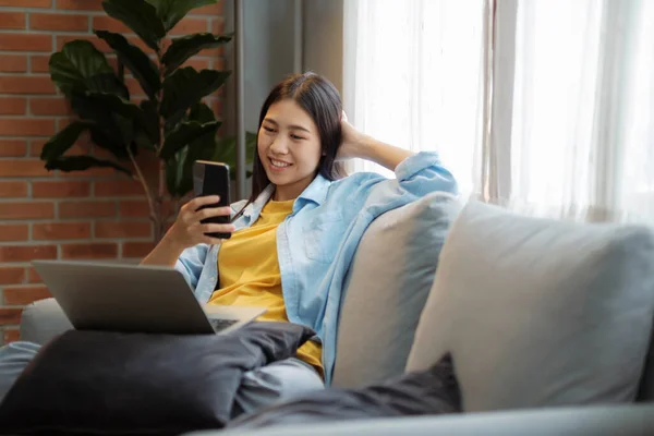 年轻的亚洲女人在家里坐在沙发上笑着用智能手机 在学习后放松 在沙发上放松时发短信或浏览社交网络 快乐的女人在手机上看视频 — 图库照片