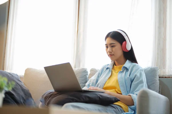 年轻的亚洲女人在家里坐在沙发上 一边学习 一边上网学习 一边用耳机听音乐 收听播客 有声书籍 使用笔记本电脑观看录像 — 图库照片