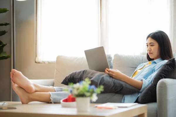 年轻的亚洲女人笑着 一边用笔记本电脑学习或工作 一边靠在沙发上 女人在明亮的客厅舒服地坐在沙发上 一边上网学习 一边用笔记本电脑搜索信息 在办公桌上学习 — 图库照片