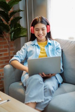 Genç Asyalı kadın çalışıyor, internetten bilgi topluyor ve evdeki kanepede otururken kulaklıkla müzik dinliyor. Podcast dinlemek, sesli kitap, gülümsemek ve dizüstü bilgisayar kullanarak video izlemek..