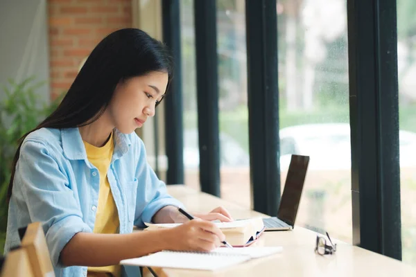 快乐的年轻亚洲女人坐在桌旁 在笔记本前写字 学生们坐在窗边的桌子边 一边用笔记本电脑学习 一边记笔记 学习概念 — 图库照片