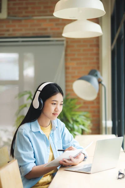 幸せな若いアジアの女性がテーブルに座ってノートパソコンを使って勉強し ノートパソコンをテーブルに座ってヘッドセットを着用してオンラインで学び 勉強します オンライン学習の概念 — ストック写真
