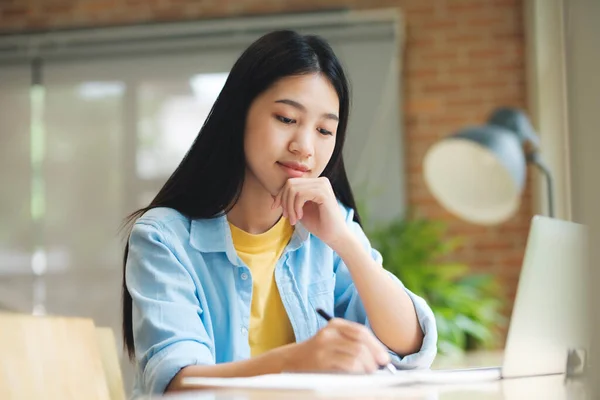 幸せな若いアジアの女性がテーブルに座ってノートパソコンの前でノートパソコンに書きます ノートパソコンで学習し 窓の横のテーブルに座りながら学校の勉強についてメモを取ります 学びの概念 — ストック写真