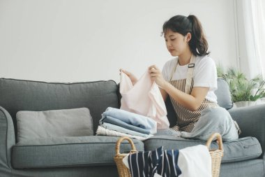 Genç bir kadın evdeki oturma odasında kanepede otururken elbiselerini katlıyor. Ev işleri yapan, çamaşır yıkayan ve evde çamaşır katlayan bir kadın. Oda servisi kavramı.
