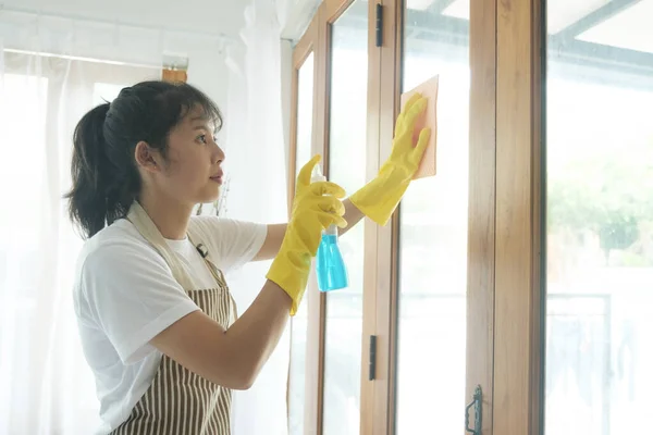窓のスプレーを使用して自宅で美しい若いアジアの女性のクリーニングウィンドウと黄色の手袋を着用 女性はそれにきれいな解決策を噴霧し 拭くことによって窓ガラスの掃除の家事を行うことを楽しむ — ストック写真