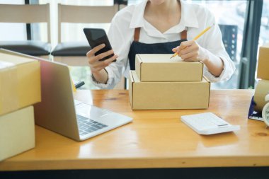 Asyalı kadın küçük bir internet şirketi ya da dükkan sahibi teslimat için cep telefonuyla alışveriş kutusunu paketliyor ve web sitesinin perakende siparişini kontrol ediyor. Çevrimiçi iş ve e-finans kavramı.
