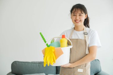 Ayakta duran ve elinde temizlik malzemeleriyle dolu bir kova tutan genç bir kadın. Ev işlerini yapmaya hazır ve mutlu bir kadın. Oda servisi kavramı.