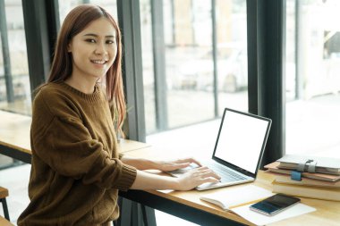 Asyalı genç kız öğrenci ya da işçi internetten ya da uzaktan öğreniyor dizüstü bilgisayarla çalışıyor ve iş yerindeki ders kitaplarıyla kameraya bakıyor ve gülümsüyor. Çevrimiçi öğrenme ve çalışma konsepti.