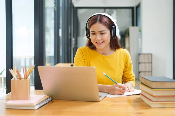 若いアジアの女性学生は オンラインコースを学び 机に座りながらノートを書き ヘッドフォンを装着したラップトップや教科書を使用して眼鏡をかけています オンライン学習と学習の概念 — ストック写真