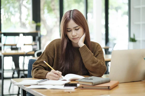 Genç Asyalı gözlüklü kız öğrenci online kuzenlerini öğreniyor ve masa başında oturup dizüstü bilgisayar ve ders kitapları kullanarak notlar yazıyor. Çevrimiçi çalışma ve öğrenme kavramı.