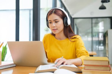 Genç Asyalı gözlüklü kız öğrenci online kuzenlerini öğreniyor ve masada otururken notlar yazıyor ve dizüstü bilgisayar ve kulaklık takıyor. Çevrimiçi çalışma ve öğrenme kavramı.