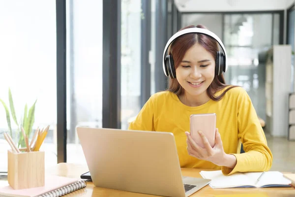若いアジアの女性学生は ノートパソコンを使ってオンラインで勉強したり 携帯電話を持ってインターネットをサーフィンしたり 机に座っている間にビデオを見たりします オンライン学習の概念 — ストック写真