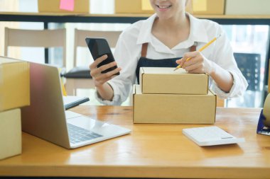 Asyalı kadın küçük bir internet şirketi ya da dükkan sahibi teslimat için cep telefonuyla alışveriş kutusunu paketliyor ve web sitesinin perakende siparişini kontrol ediyor. Çevrimiçi iş ve e-finans kavramı.