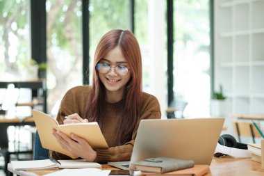 Genç Asyalı gözlüklü kız öğrenci online kuzenlerini öğreniyor ve masa başında oturup dizüstü bilgisayar ve ders kitapları kullanarak notlar yazıyor. Çevrimiçi çalışma ve öğrenme kavramı.