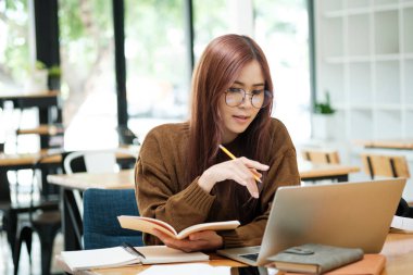 Genç Asyalı gözlüklü kız öğrenci online kuzenlerini öğreniyor ve masa başında oturup dizüstü bilgisayar ve ders kitapları kullanarak notlar yazıyor. Çevrimiçi çalışma ve öğrenme kavramı.