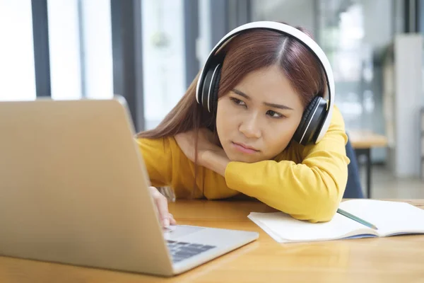 Young female student or worker using laptop to study or work online and feeling bored, tired, and disappointed while sitting and leaning on her desk. Online learning or work concept.