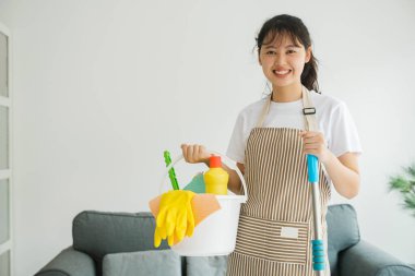 Ayakta duran ve elinde temizlik malzemeleriyle dolu bir kova tutan genç bir kadın. Ev işlerini yapmaya hazır ve mutlu bir kadın. Oda servisi kavramı.