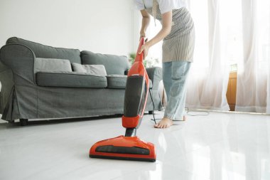 Günlük kıyafetlerle yerleri temizleyen, elektrikli süpürge kullanan ve evdeki temizlik malzemelerini kullanan genç bir kadın. Oda servisi kavramı.