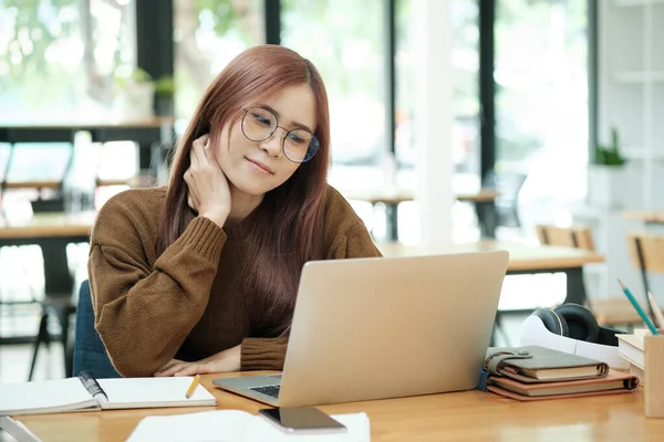 Young asian female student or worker learning or working online using laptop and giving a confused face and gesture while sitting at dask. Online work or learning concept.