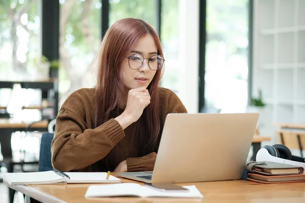Asian young female student or worker learning online or distant working using laptop and sitting at desk with textbooks and stationaries lying at workplace. Online learning and working concept.