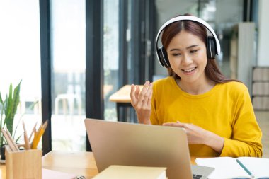 Genç Asyalı kız öğrenci internetten okuyor ve öğreniyor. İş yerinde masa başında otururken kulaklık takarak bilgisayarla video görüşmesi yapıyor. Çevrimiçi öğrenme kavramı.