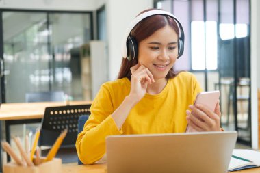 Genç Asyalı kız öğrenci dizüstü bilgisayar kullanıyor ve cep telefonu tutuyor bilgi almak için internette sörf yapıyor ya da Dask 'ta otururken video izliyor. Çevrimiçi öğrenme kavramı.