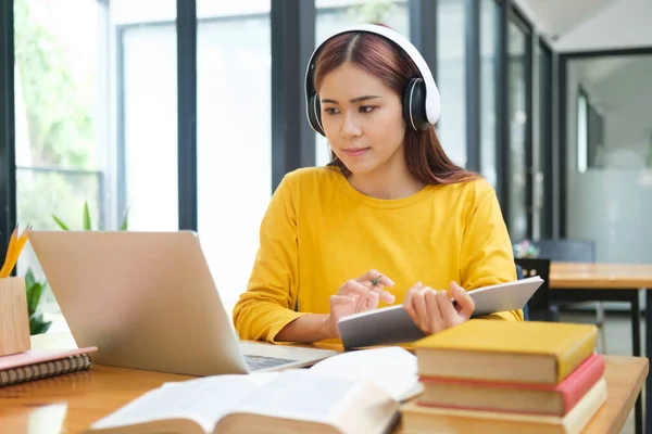 戴眼镜的年轻亚洲女学生坐在办公桌前 戴着耳机 使用笔记本电脑和课本 一边学习网上婚纱 一边写笔记 在线学习和学习概念 — 图库照片