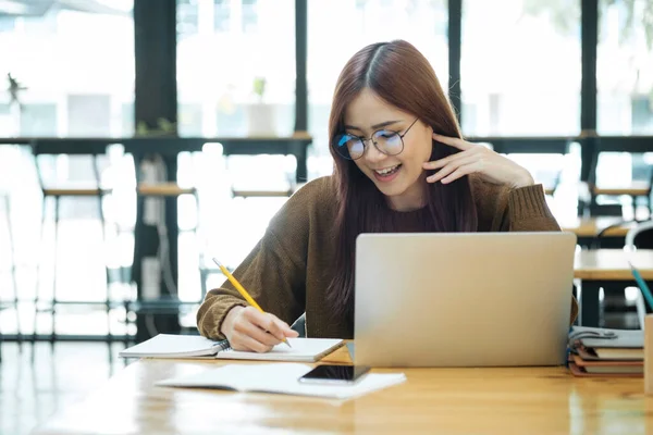 戴眼镜的年轻亚洲女学生坐在办公桌前 使用笔记本电脑和课本 一边学习网上婚纱 一边写笔记 在线学习和学习概念 — 图库照片