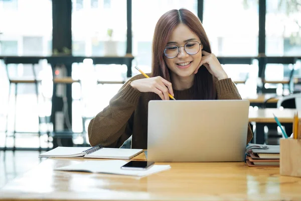 Asian young female student or worker learning online or distant working using laptop and sitting at desk with textbooks and stationaries lying at workplace. Online learning and working concept.