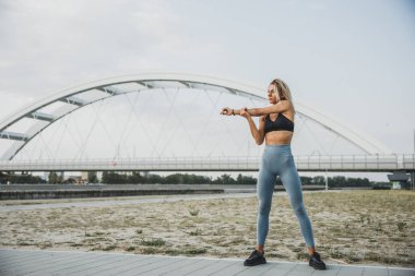 Formda genç bir kadın ısınıyor ve kollarını nehir köprüsünün yanında geriyor..