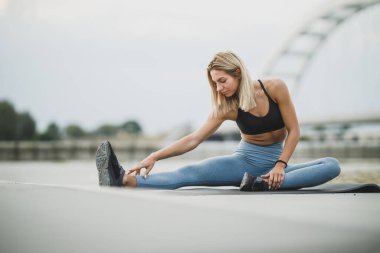 Nehir köprüsü yakınlarında spor antrenmanı için ısınırken esneme egzersizleri yapan genç bir kadın..