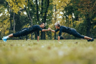 Formda bir çift, kaslarını canlandırmak için siyah takım elbiseli ve EMS elektronik simülatörlü parkta tahta egzersizleri yapıyor..