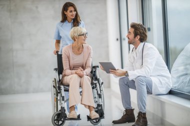 Başarılı doktor ve genç hemşire tekerlekli sandalyeye mahkum yaşlı kadın hastalarıyla konuşuyor..