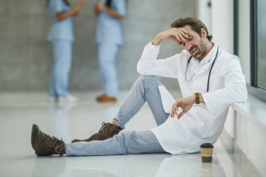 Yorgun ve olgun bir doktor, Covid-19 salgını sırasında boş bir hastane koridorunda kahve molası verirken pencerenin yanında oturup saatine bakıyor..